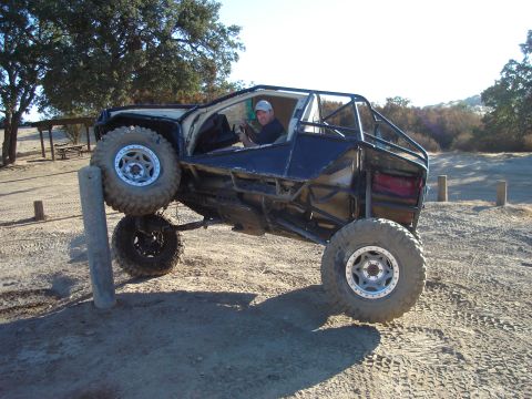 1985 Toyota Hilux - Custom off-road buggy with large tires navigates an obstacle on a dirt track.