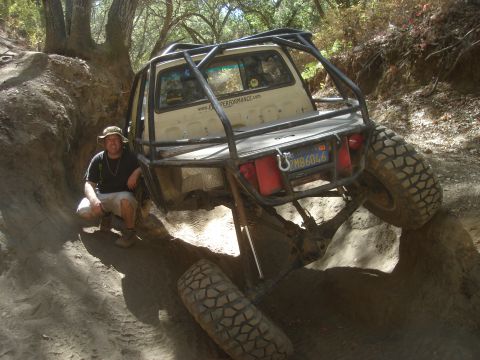 1985 Toyota Hilux - A man poses next to a custom off-road truck navigating a steep, muddy incline.