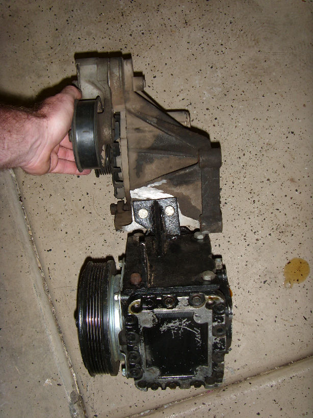 Hand holding an AC compressor and mounting bracket, resting on concrete floor