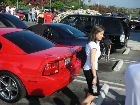 1998 Ford Crown Victoria - A red Ford Mustang is parked at a car meet, with other vehicles and people in the background.
