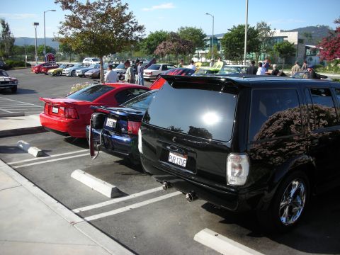 1998 Ford Crown Victoria - A black SUV with custom wheels and a red Mustang are parked side-by-side in a sunny parking lot.