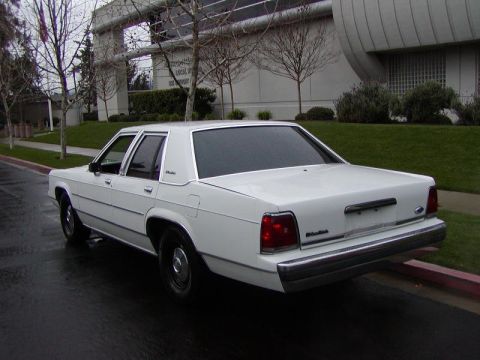 1991 Ford LTD Crown Victoria - The rear quarter panel of a white sedan is visible, parked on a wet street next to a curb.