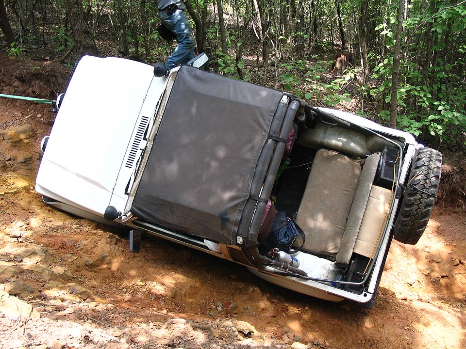 White 4x4 SUV rolled onto its side on a dirt trail in the woods, occupant standing on it