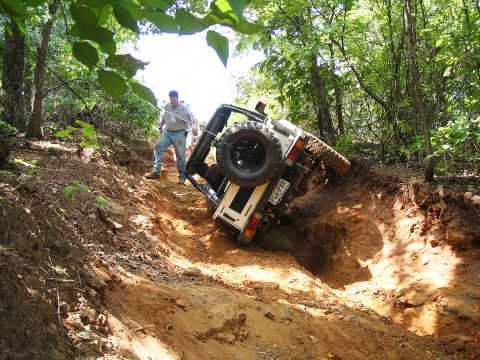 1987 Suzuki Samurai - A person attempts to pull a white off-road vehicle out of a steep ditch where it has become stuck on its side.