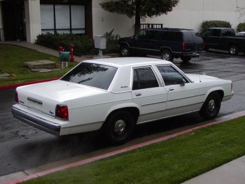 1991 Ford LTD Crown Victoria - Rear three-quarter view of a clean white Ford Crown Victoria four-door sedan parked at a red-painted curb on wet pavement outside a commerc…