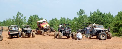 1987 Suzuki Samurai - Several off-road vehicles are gathered on a dirt track with trees and a clear sky overhead.