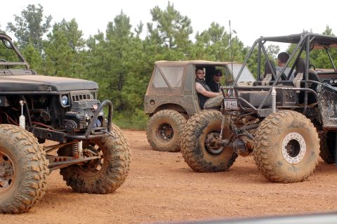 1987 Suzuki Samurai - Several heavily modified off-road vehicles are parked on a dirt path surrounded by trees.