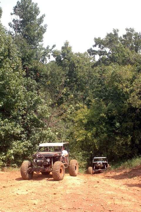 1987 Suzuki Samurai - Two off-road buggies are seen on a dirt path surrounded by trees.
