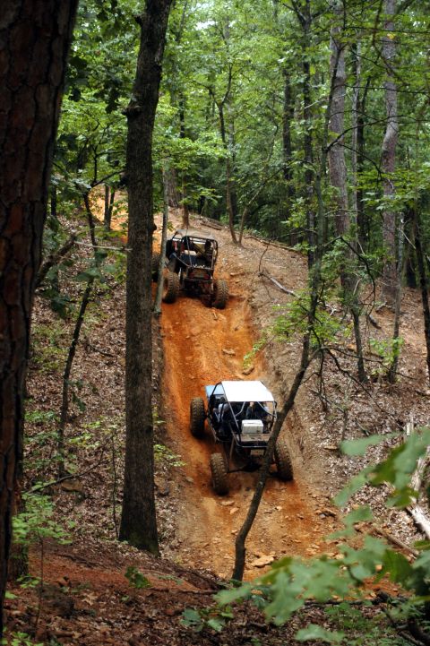 1987 Suzuki Samurai - Two custom off-road vehicles navigate a challenging dirt track through a wooded area.