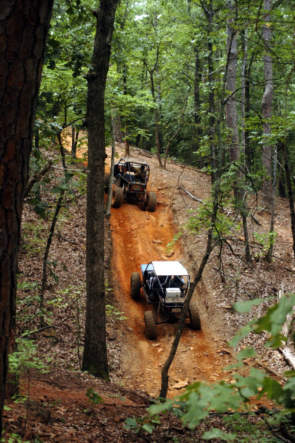 Two off-road buggies ascend a steep, muddy trail in a dense forest