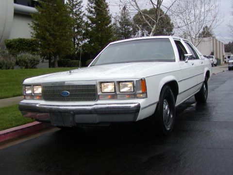 1991 Ford LTD Crown Victoria - White Ford Crown Victoria sedan with headlights on, parked at a curb on wet pavement and shot low from the 3/4 front left. A whip antenna i…