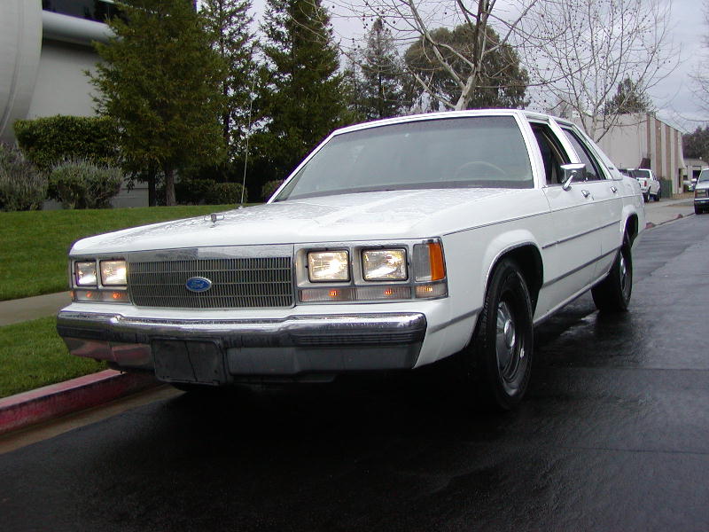 White Ford Crown Victoria sedan with headlights on and fender whip antenna, wet street