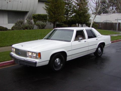 1991 Ford LTD Crown Victoria - Front three-quarter view of a clean white Ford Crown Victoria four-door sedan on wet pavement beside a manicured lawn and hedge, headlights…