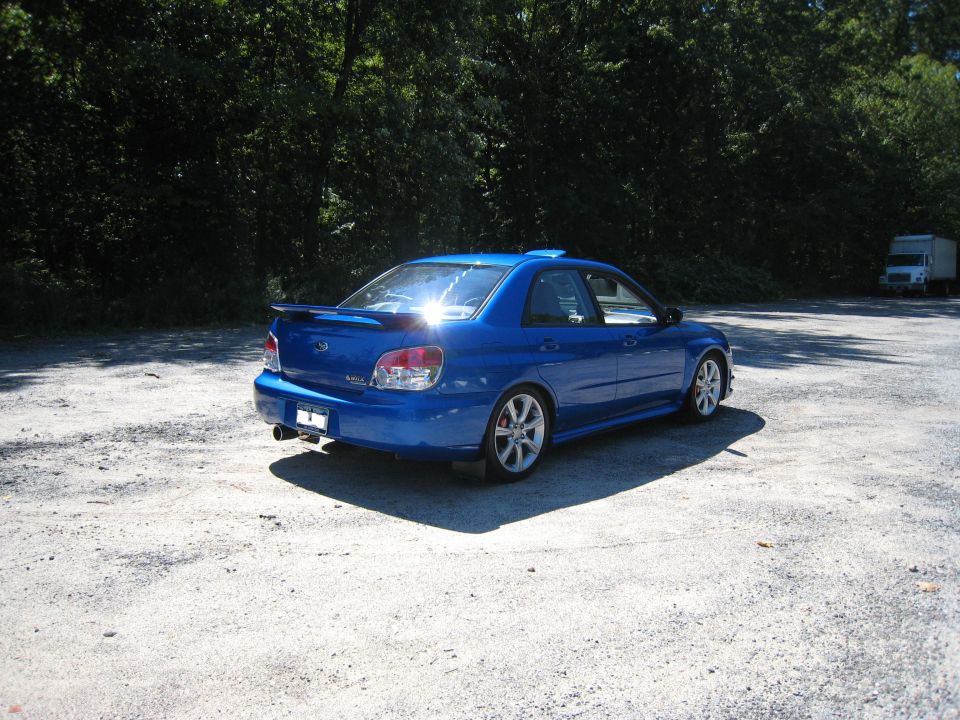 Blue Subaru Impreza sedan, rear 3/4 view, parked on gravel lot with trees behind