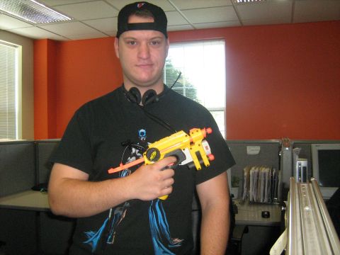 1988 Mercury Grand Marquis - A man poses with a toy gun in an office environment, with cubicles and computers visible.