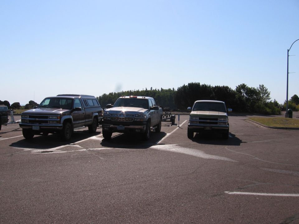 Three pickup trucks parked in a lot under a clear sky. The truck on the left is dark brown with a camper shell. The middle truck is silver and appears newer. The truck on the right is light tan