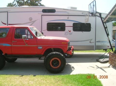 1985 Ford Bronco - A red SUV with off-road tires is parked next to a large RV, with a basketball hoop in the background.