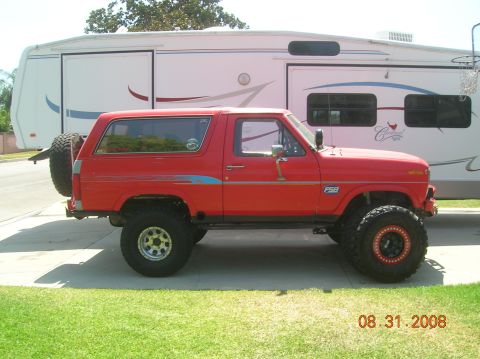 1985 Ford Bronco - A modified red Ford Bronco is parked on a driveway, with a large RV behind it.