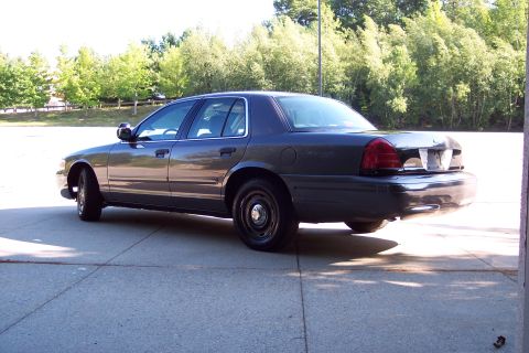 2001 Ford Crown Victoria - A dark gray sedan is viewed from the rear quarter angle, parked outdoors on a concrete surface with trees in the background.