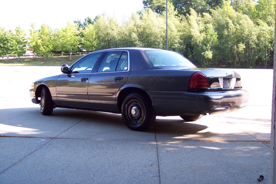 Dark gray sedan parked on concrete, rear quarter view. Black steel wheels, tinted windows, and a police-style antenna visible