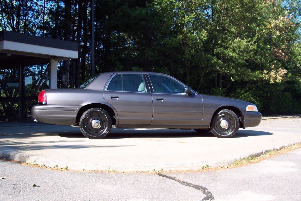 Gray Ford Crown Victoria parked on concrete. Black steel wheels. Trees and building in background