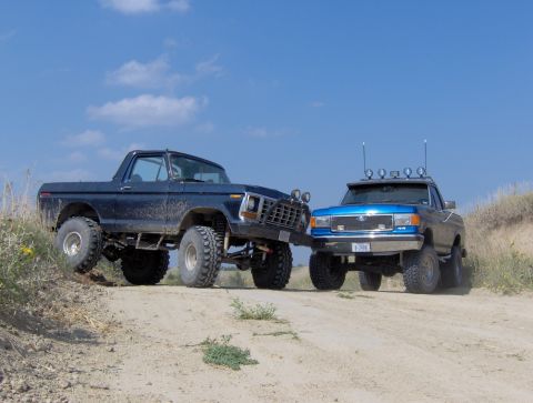 1987 Ford Bronco - A blue 1987 Ford F-Series truck is parked next to a dark-colored older model truck on a dirt road under a blue sky.
