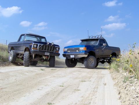 1987 Ford Bronco - Two lifted Ford pickup trucks are parked on a dusty dirt road under a blue sky.