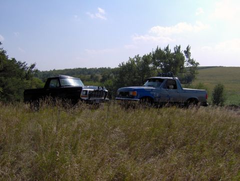 1987 Ford Bronco - Two pickup trucks are parked in a field with trees and hills in the background under a partly cloudy sky.