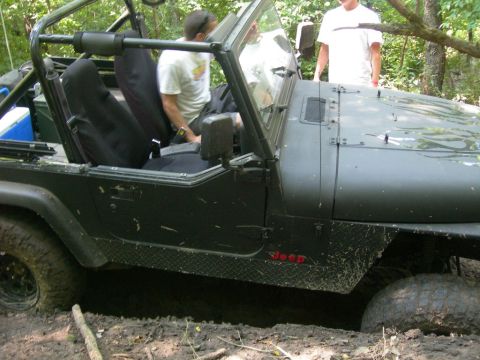 1991 Jeep Wrangler - A dark green Jeep Wrangler is stuck in the mud, with two people visible inside and standing nearby.