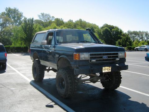 1990 Ford Bronco - Front three-quarter view of a lifted dark blue Ford Bronco with a white removable hardtop, parked in a sunny asphalt lot on oversized mud-t…