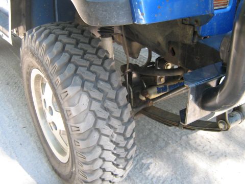 1990 Ford Bronco - Close-up view of the front suspension and a large off-road tire on a blue vehicle. The "Supermotors.net" watermark is visible.