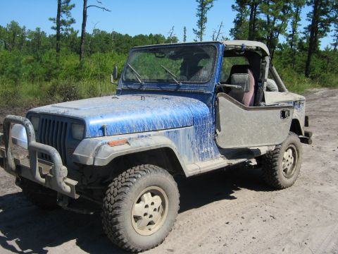 1990 Ford Bronco - A dusty blue Jeep Wrangler sits on a dirt road, its driver's side door ajar, ready for off-road adventure.