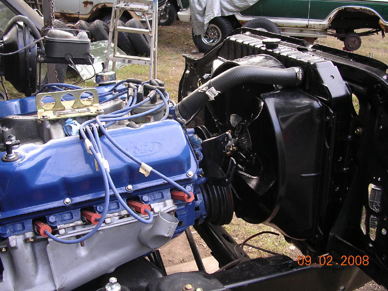 Ford V8 engine with blue valve covers and spark plug wires. Radiator and fan visible. Other vehicles in background