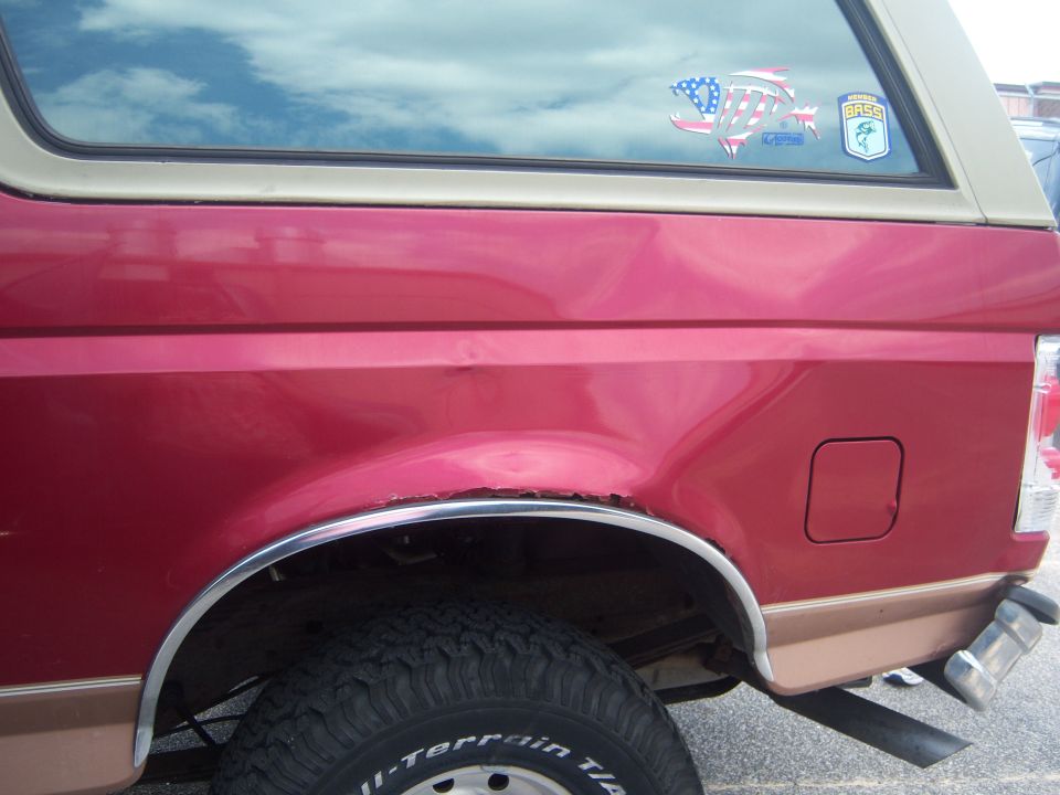 Red and tan Ford Bronco rear quarter panel with dents and rust, showing a tire and fuel door