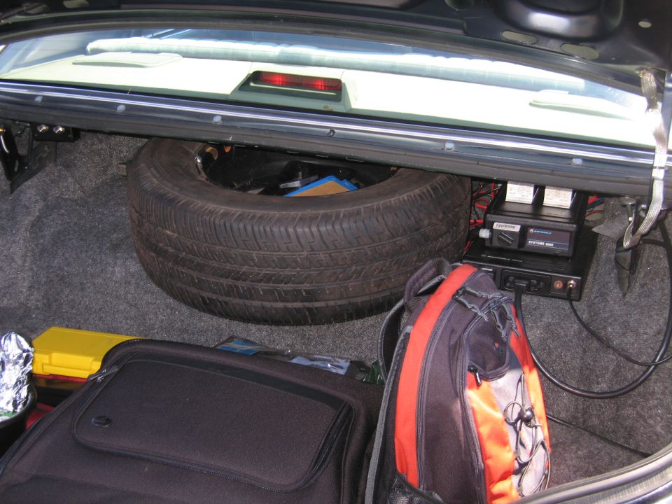 Car trunk interior with spare tire, black suitcase, orange and black backpack, and radio equipment mounted on the side