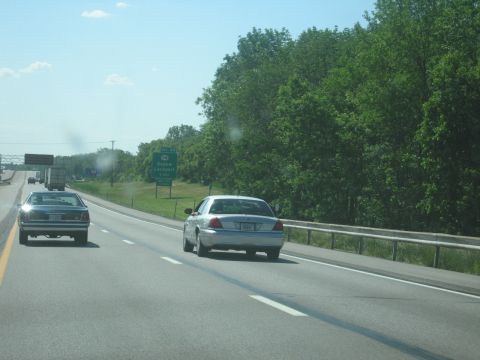 2001 Ford Crown Victoria - Two cars driving on a highway with trees and a green sign in the background.