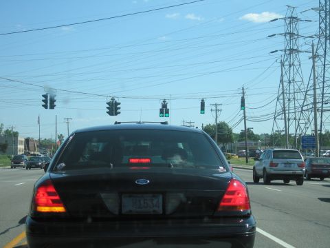 2001 Ford Crown Victoria - Rear view of a black 2001 Ford Crown Victoria driving on a sunny day, with traffic lights and power lines overhead.