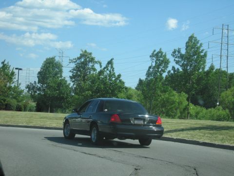 2001 Ford Crown Victoria - A dark sedan is seen from behind while driving on a paved road under a partly cloudy sky.