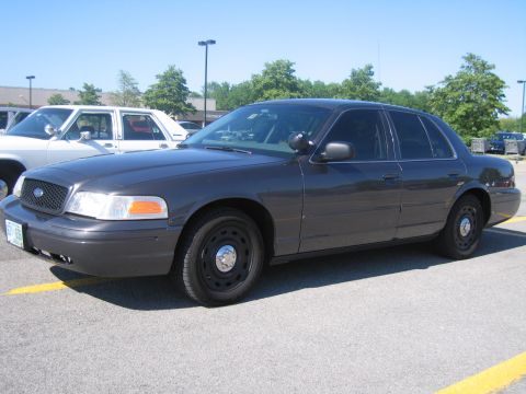 2001 Ford Crown Victoria - A dark sedan, possibly a 2001 Ford Crown Victoria, is parked in a lot next to a white vehicle under a clear sky.