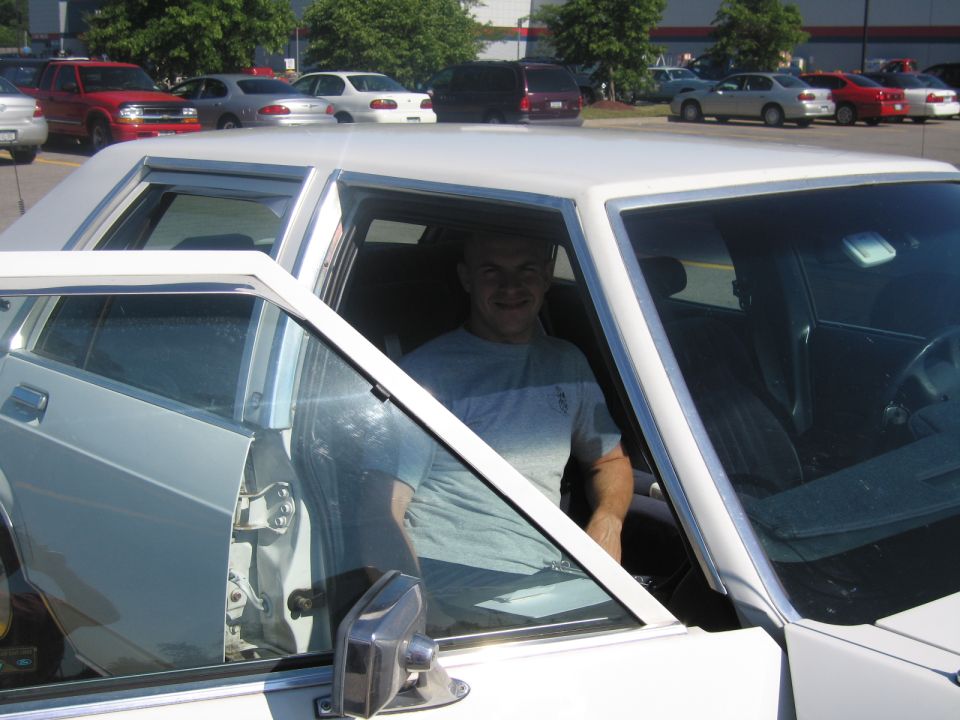 man in driver's seat of white sedan, passenger door open; other cars in background parking lot