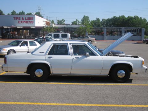 2001 Ford Crown Victoria - A white sedan is parked in a lot with the hood open, showing other vehicles and a building in the background.