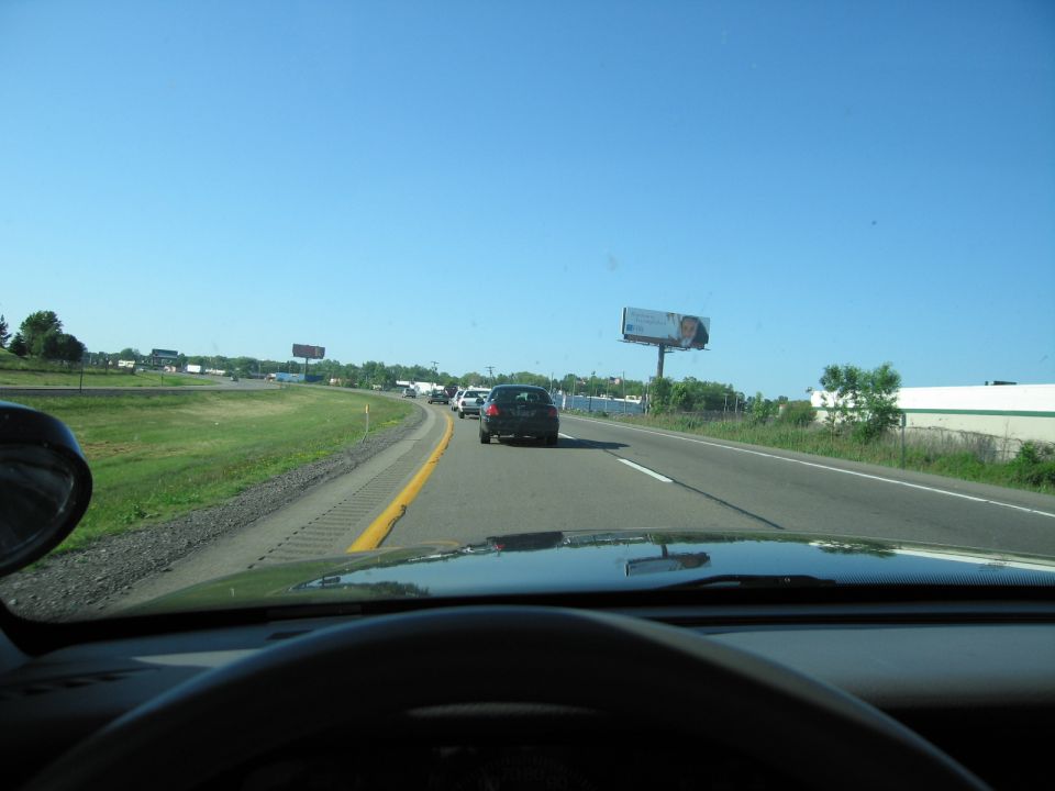 View from inside a car driving on a highway with other cars ahead and billboards on the side