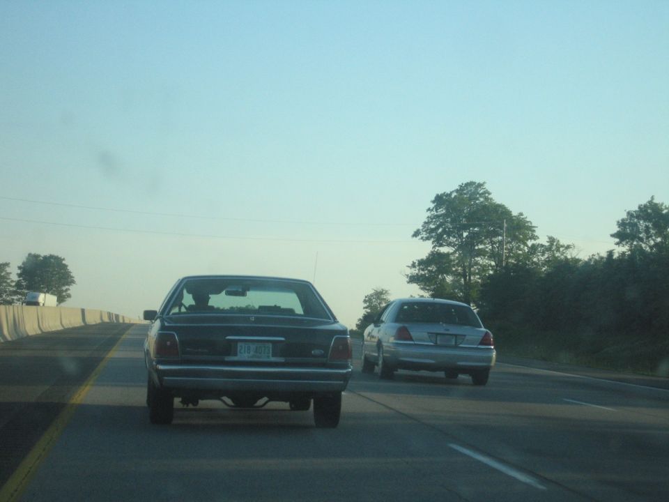 Two sedans, one dark and one silver, driving on a highway. The dark sedan is in the foreground, and the silver sedan is ahead