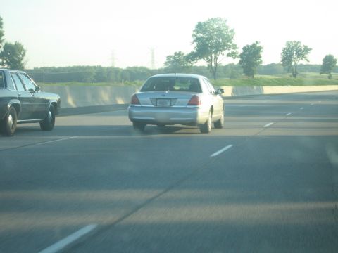 2001 Ford Crown Victoria - Two cars travel down a highway under a hazy sky with trees and power lines in the background.