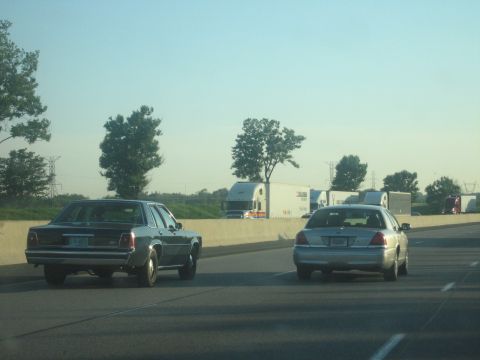 2001 Ford Crown Victoria - Two sedans travel on a highway under a clear blue sky, with trucks visible in the background.