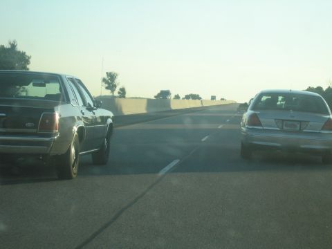 2001 Ford Crown Victoria - Two sedans, one silver and one dark, travel on a highway in what appears to be a 2001 Ford Crown Victoria.