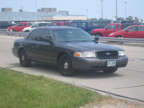 2001 Ford Crown Victoria - A dark grey 2001 Ford Crown Victoria sedan is parked on the side of the road with other vehicles in the background.