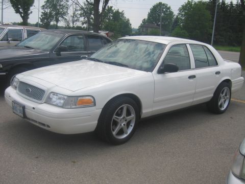 2001 Ford Crown Victoria - A white 2001 Ford Crown Victoria sedan is parked in a lot, showing its front and side profile. Another dark sedan is visible behind it.