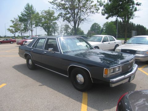 2001 Ford Crown Victoria - A dark sedan is parked in a lot with other vehicles visible in the background under a partly cloudy sky.