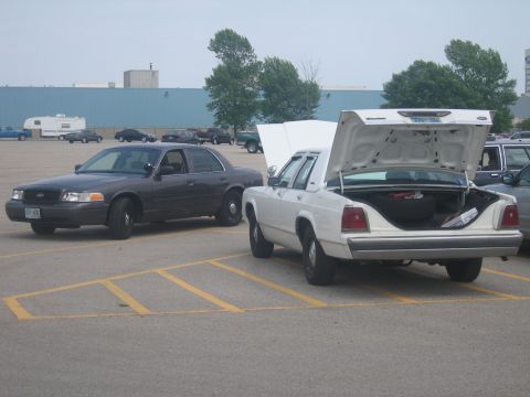 2001 Ford Crown Victoria - Two Ford Crown Victorias are parked in a lot, one white with its trunk and hood open, the other dark grey.